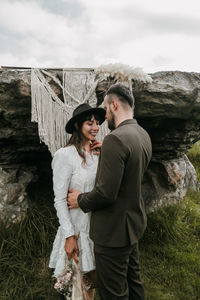 Delighted newlywed couple in stylish clothes standing against macrame hanging on rock in nature on wedding day and embracing