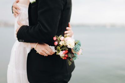 Midsection of newlywed couple embracing while standing by sea against sky