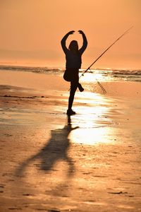 Full length of silhouette teenage girl with arms raised standing on one leg at beach during sunset