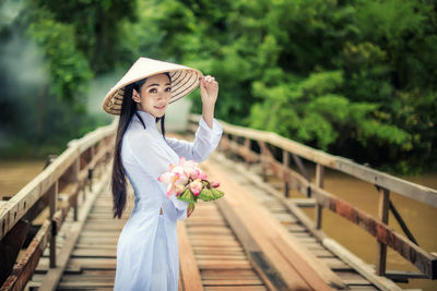Portrait of young woman wearing conical hat while holding flowers on footbridge