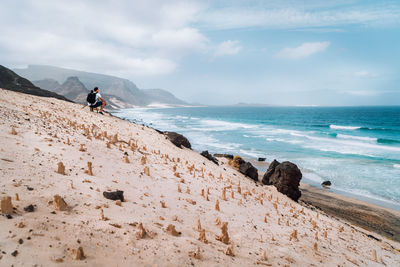 Man crouching on hill at beach against sky