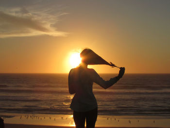 Rear view of carefree young woman removing rubber band from hair while standing on beach against sky during sunset