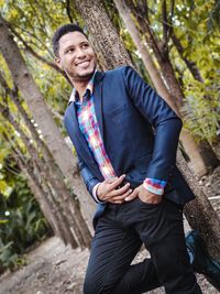 Smiling young man in suit looking away while standing against tree