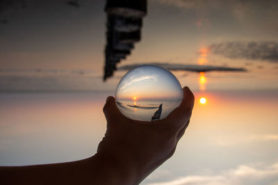 Cropped hand holding crystal ball with reflection of sea during sunset
