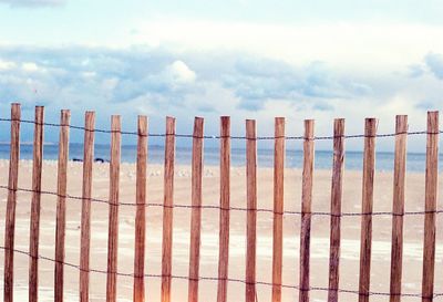 Wooden fence at beach against cloudy sky