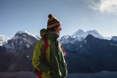 Hiker in warm clothing looking away while standing on mountain against blue sky