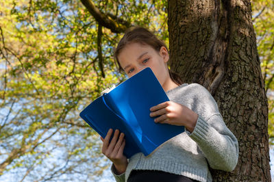 Young school age girl reading from blue textbook in park leaning against tree.