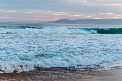 Waves rushing towards shore against sky during sunset