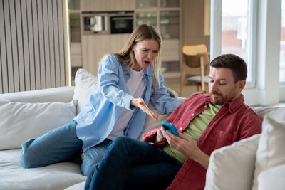 Upset emotional annoyed wife shouting at indifferent husband sitting on sofa, busy with mobile phone