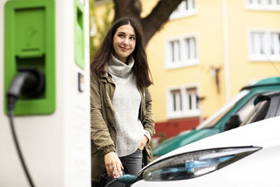 Smiling beautiful woman looking away while standing with hand in pocket and charging electric vehicle at station