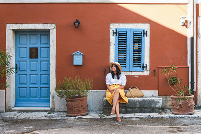 Young woman in summer outfit sitting on bench in front of colorful house.