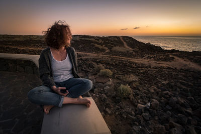 Woman holding mobile phone while sitting outdoors at sunset