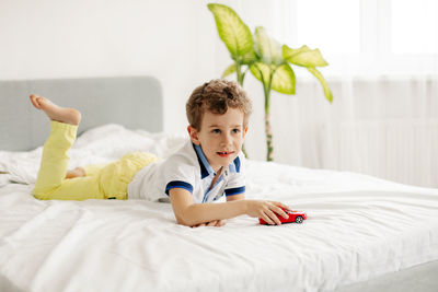 A little boy is lying on his stomach and playing with a toy car on his bed