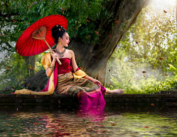 Young woman with umbrella sitting by lake in forest