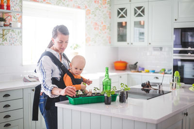 Working mother planting plants in container while carrying daughter in kitchen at home