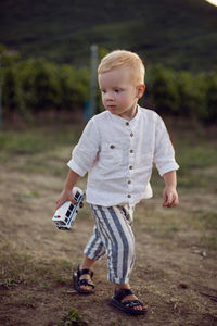 Blond boy in striped trousers stands in a vineyard at sunset holding a vine