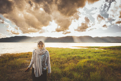 Smiling woman wearing headscarf while standing on grassy field against cloudy sky