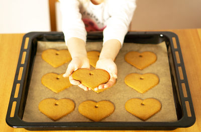 Girl hands with a heart cookie in front of the homemade fresh baked cookies on baking sheet.