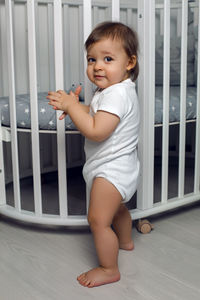 Child in white clothes of one year stands on the floor next to his round white bed