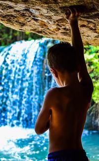 Rear view of shirtless boy standing against waterfall