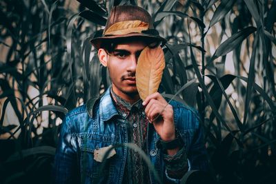 Portrait of young man holding leaf while standing by plants