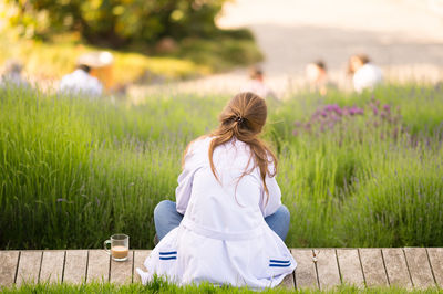 Rear view of woman sitting by coffee cup on boardwalk against grass