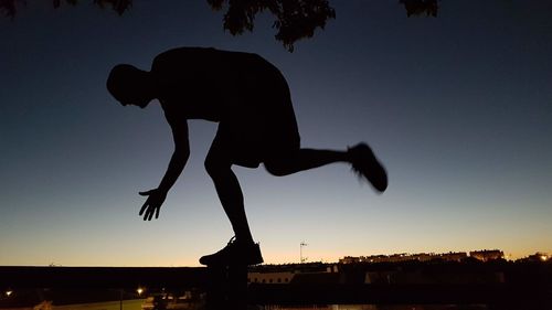 Optical illusion of silhouette man jumping on city against clear sky during sunset