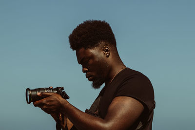 Side view of young man photographing against clear blue sky