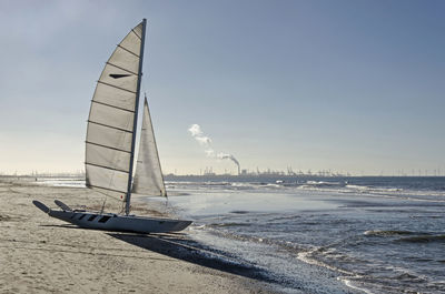 Catamaran sailboat on an almost empty sandy beach , with the port of rotterdam in the background