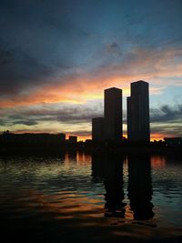 Silhouette buildings by lake against sky during sunset