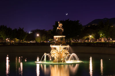 Illuminated fountain by lake against sky at night