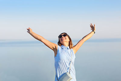 Cheerful woman with arms raised standing against sea