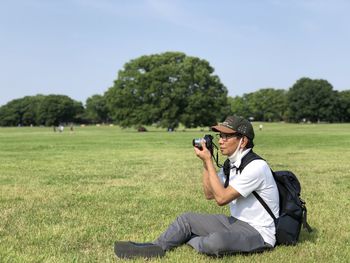 Side view of man photographing on field behind big tree background garden.