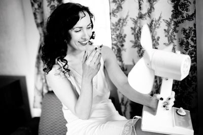 Smiling young woman holding electric fan while smoking cigarette at home