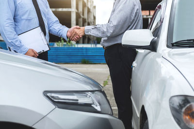 Midsection of insurance agent shaking hands with customer while standing by cars outdoors