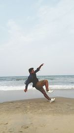 Side view of young man jumping on shore at beach against sky