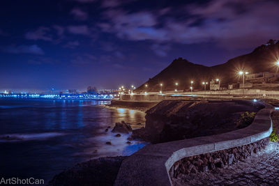 Illuminated city by sea against sky at night