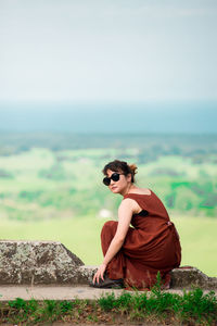 Portrait of young woman wearing sunglasses sitting on observation point against sky