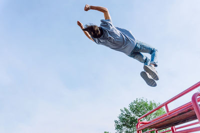 From below of brave male jumping over metal fence in street and showing parkour trick against blue sky