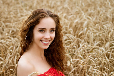 Portrait of a young cheerful woman with wavy hair in a red dress sits in a wheat field