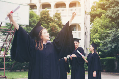 Cheerful student in graduation gown with arms raised standing outdoors