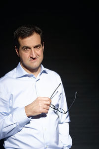 Portrait of man holding eyeglasses against black background
