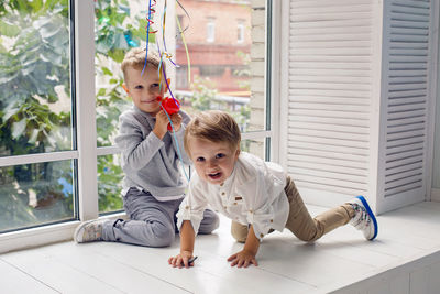 Two brothers sitting by the window in a white studio with green trees on the street