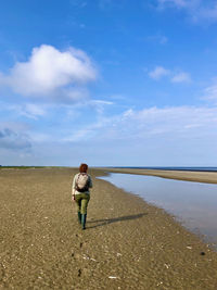 Rear view of woman walking along an empty shore under a blue sky with white clouds