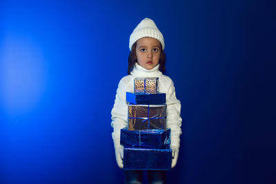 Christmas boy child in a white knitted sweater and hat stands in the studio against gifts boxes