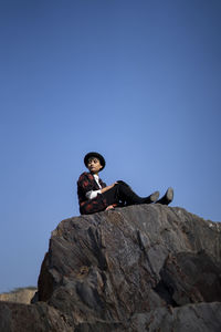 Low angle view of man sitting on rock against clear blue sky