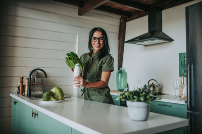 Portrait of woman holding bottle and vegetable while standing at home