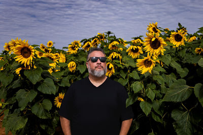 Portrait of young man standing amidst sunflowers against sky