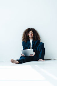 Businesswoman with digital tablet wearing oversized suit while sitting in front of wall