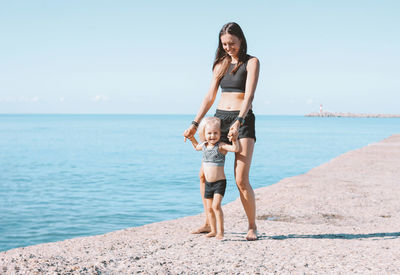 Full length of mother and daughter standing at beach against sky